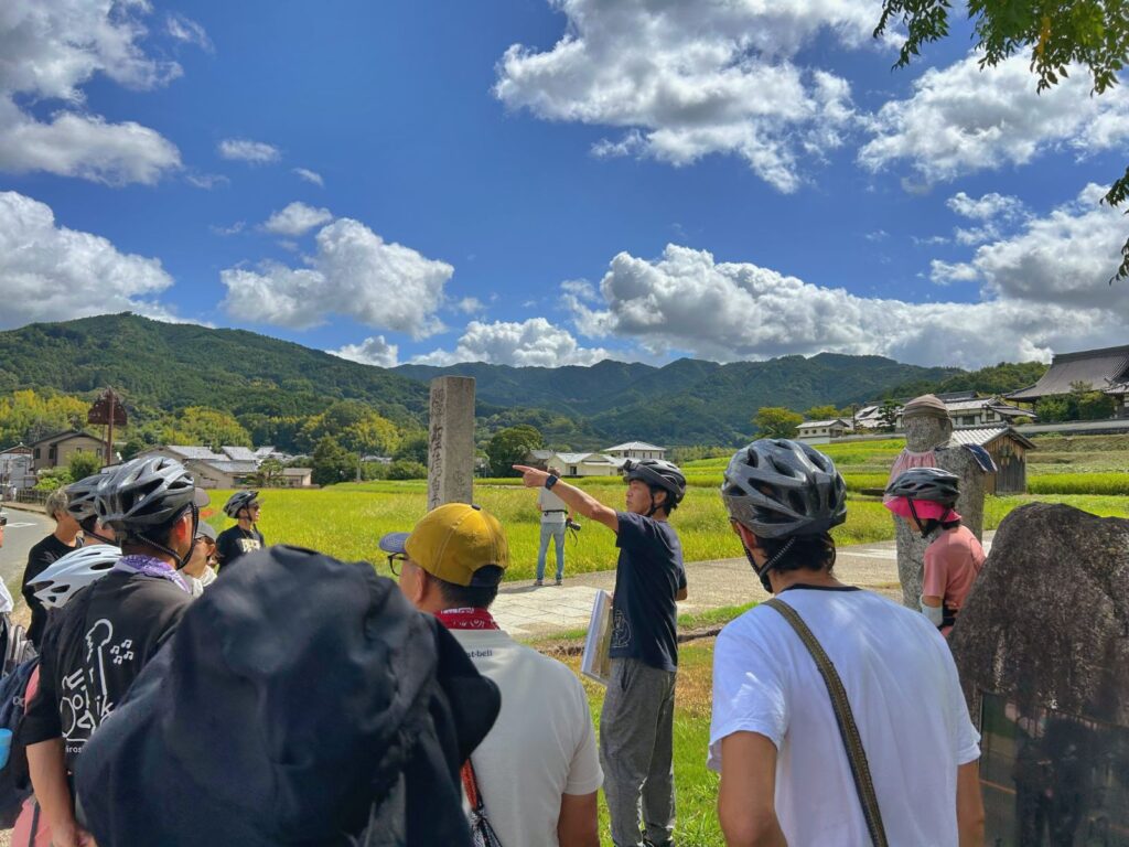 Travelers listening to a local guide explaining Asuka’s history beside rice fields and mountains